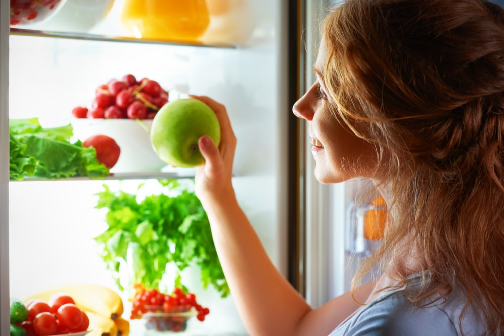 Woman holding an apple looking into an open refrigerator